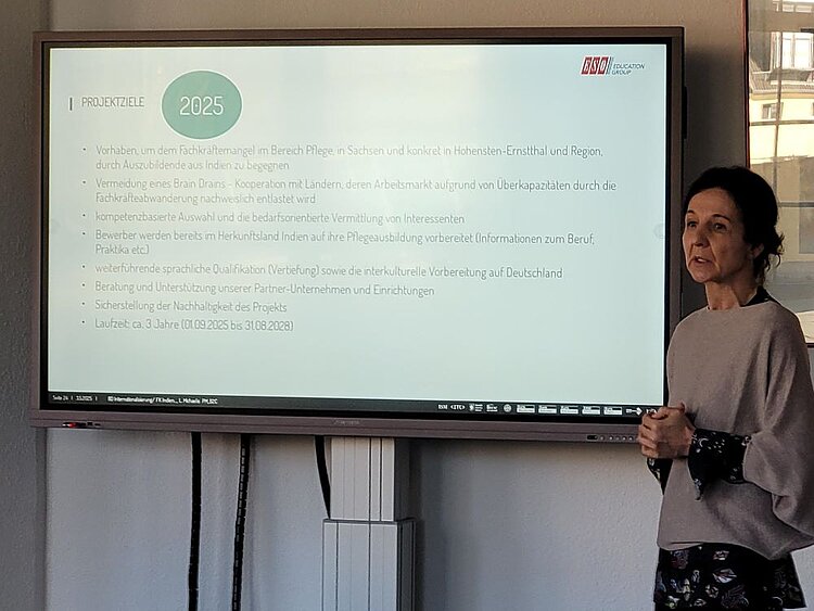 Eine Frau steht neben einem großen Bildschirm und hält eine Präsentation. Auf dem Bildschirm sind die „Projektziele 2025“ einer Initiative zur Bekämpfung des Fachkräftemangels in der Pflegebranche in Deutschland durch die Rekrutierung von Auszubildenden aus Indien dargestellt. Die Präsentationsfolie enthält Punkte zu Themen wie Vermeidung von „Brain Drain“, sprachliche Qualifikation, interkulturelle Vorbereitung und Nachhaltigkeit des Projekts. Das Logo der „ESO Education Group“ ist in der oberen rechten Ecke zu sehen. Die Frau trägt eine beige Strickjacke über einem dunklen Kleid mit Blumenmuster und spricht zur Gruppe.