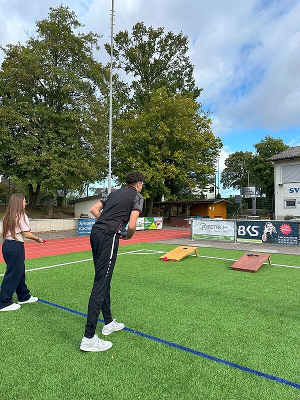 Together in motion – Bewegung, Spaß und Teamgeist Zwei Personen spielen auf einem Sandplatz Cornhole. Eine Person hält einen Beanbag in der Hand und zielt auf eines der beiden Spielbretter. Im Hintergrund sind Bäume und ein Sportplatz zu sehen.