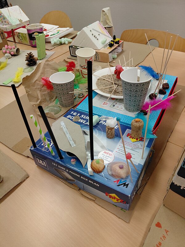 Auf einer blauen Technik-Verpackung ist eine Konstruktion aus Pappbechern, Holzspießen, Korken, Muscheln und bunten Federn aufgebaut, die wie ein kleines Experimentierfeld oder Parcours wirkt; im Hintergrund sind weitere Bastelwerke sichtbar.