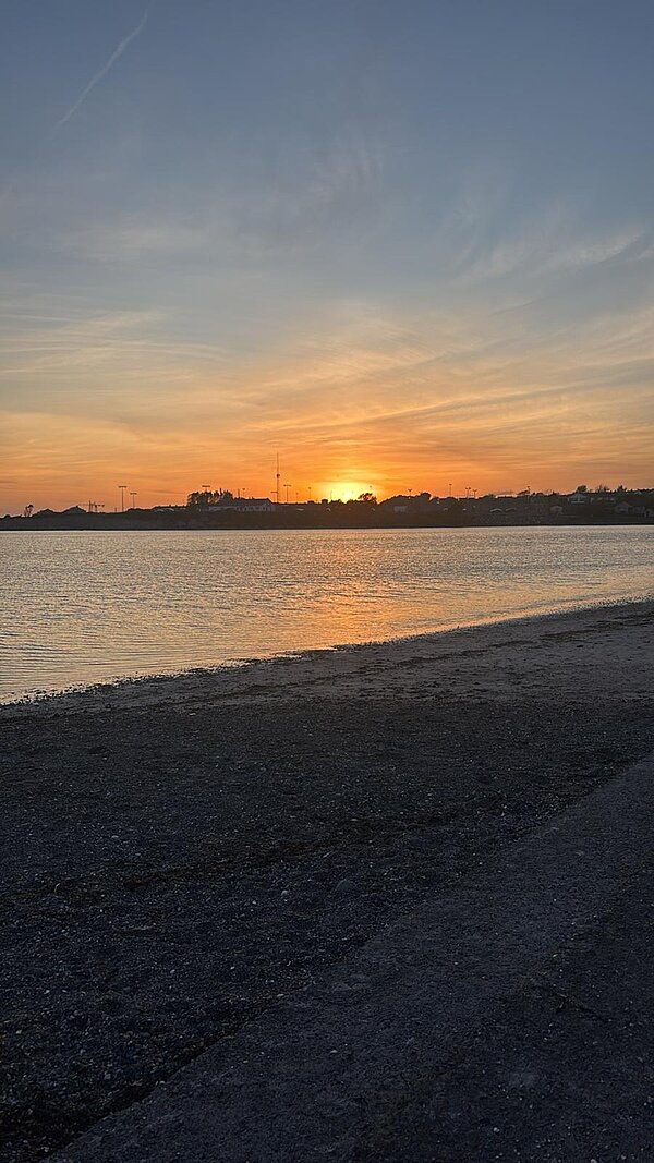 Blick vom kieseligen Strand auf die untergehende Sonne und die Skyline. 