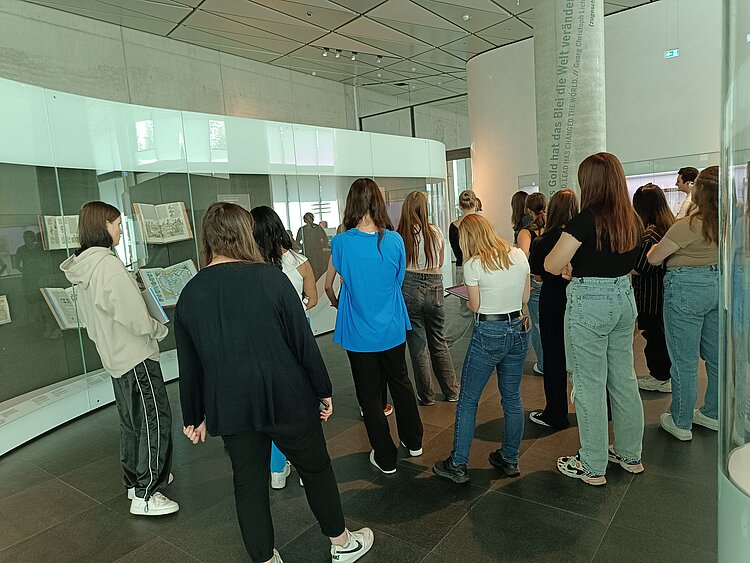 Junge Frauen stehen in einem Halbkreis um eine Person, die ihnen etwas erklärt. Die Frauen sind mit Jeans, schwarzen Hosen, dunklen und blauen Oberteilen oder grauen Hoodies bekleidet.