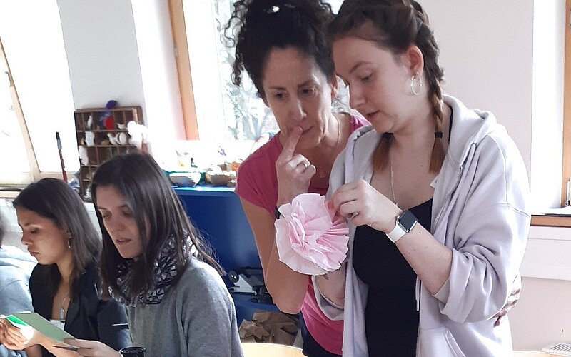 Vier Personen sitzen und stehen an einem Tisch, auf dem Bastelmaterialien liegen. Eine der stehenden Personen scheint der anderen, mit einer Bastelarbeit in den Händen, etwas zu erklären.