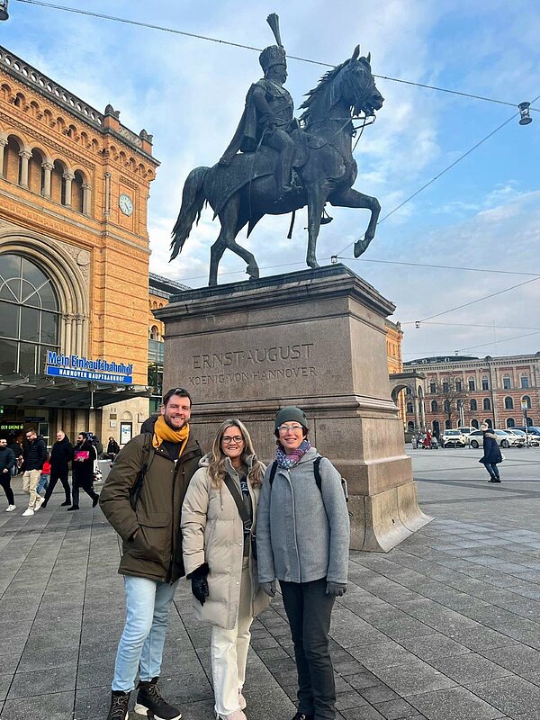 Drei Personen stehen vor dem Reiterstandbild von Ernst August, König von Hannover, das sich vor dem Hauptbahnhof Hannover befindet. Die Personen tragen Winterkleidung und lächeln in die Kamera. Im Hintergrund sind Teile des Bahnhofsgebäudes und weitere Passanten zu sehen.
