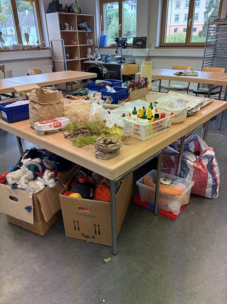 Neues Kreativprojekt: Komm, wir bauen eine Stadt! Ein großer Tisch in einem hellen Klassen- oder Bastelraum ist mit verschiedenen Materialien für kreative Aktivitäten bedeckt. Zu sehen sind Wolle, Bastelscheren, Klebstoffe, Pinsel, Naturmaterialien wie Tannenzapfen und Stroh, sowie Behälter mit weiteren Bastelutensilien. Unter dem Tisch stehen Kartons und Plastikboxen mit zusätzlichen Materialien. Im Hintergrund sind Regale, Stühle und Fenster zu erkennen.