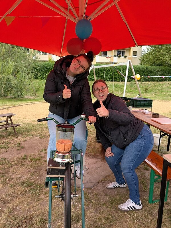 Zwei Frauen präsentieren gut gelaunt einen Fahrrad-Mixer, mit dem ein frischer Smoothie zubereitet wird.