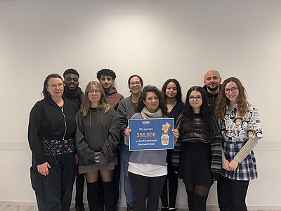 Eine Gruppe von jungen Männern und Frauen steht vor einen weißen Wand. Eine Frau hält einen blauen Spendenscheck mit Euro Akademie-Logo und der Zahl 356,00 Euro in die Kamera.