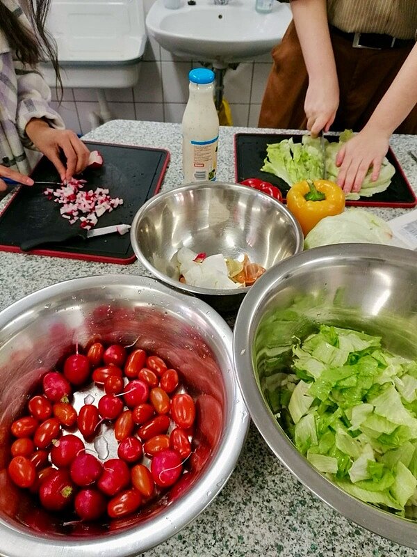 An die Töpfe, fertig, los! Auszubildende schneiden Radischen, Salat, Tomaten und Paprika, die in silbernen Schüsseln liegen.
