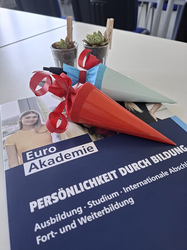Auf einem Tisch liegt eine Mappe der Euro Akademie mit dem Leitmotto "Persönlichkeit durch Bildung", darauf zwei kleine Schultütchen in rot und hellblau. Dahinter wurden zwei kleine Gläser mit Sukkulenten positioniert. Das Bild fasst den Anlass "Ausbildungsstart in Hohenstein-Ernstthal" gut zusammen.