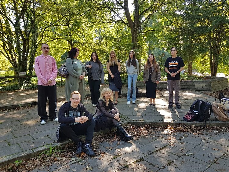 Die Gruppe der Auszubildenden steht und sitzt auf einem gepflasterten Platz im Freien. Im Hintergrund sind Bäume mit grünem Laub zu sehen, durch die Sonnenlicht fällt. Einige Azubis stehen, während zwei vorne auf dem Boden sitzen. Die Kleidung ist unterschiedlich, von leger bis formell. Auf dem Boden liegen einige Taschen und Rucksäcke.