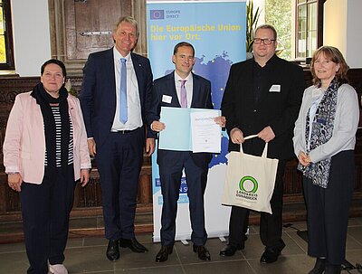 Mehrere Politiker*innen und der Schulleiter Ludwig Knapp haben sich für ein Foto positioniert, dabei wir die Urkunde "Europaschule in Niedersachsen" übergeben. Die Szene spielt sich im Osnabrücker Rathaus ab, hinter den Personen steht ein Rollup, das für die Europäische Union wirbt.