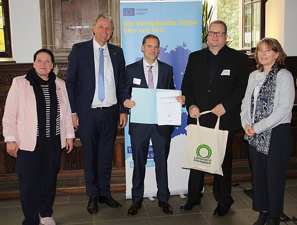 Mehrere Politiker*innen und der Schulleiter Ludwig Knapp haben sich für ein Foto positioniert, dabei wir die Urkunde "Europaschule in Niedersachsen" übergeben. Die Szene spielt sich im Osnabrücker Rathaus ab, hinter den Personen steht ein Rollup, das für die Europäische Union wirbt.