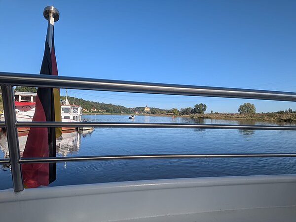 Blick von einem Schiff über einen breiten Fluss bei sonnigem Wetter. Links weht eine Deutschlandflagge am Geländer, auf dem Wasser liegen weitere Boote und am gegenüberliegenden Ufer sind Bäume und Gebäude zu sehen.