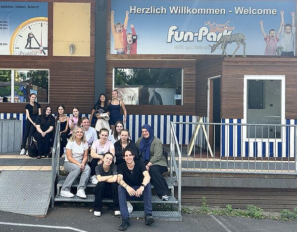 Eine Gruppe junger Menschen auf einer Treppe vor einem Gebäude mit Holzvertäfelung und blau-weißen Streifen. Oben am Gebäude hängt ein Banner mit der Aufschrift Herzlich Willkommen - Welcome - Fun-Park Würzburg. Auf dem Banner sind Tiere und jubelnde Kinder abgebildet.
