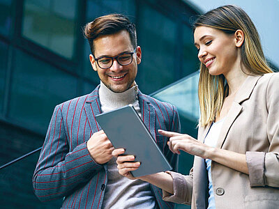 Ein junger Mann und eine junge Frau in geschäftlicher Kleidung stehen im Freien vor einem modernen Gebäude und schauen auf ein Tablet, welches die Frau hält und darauf zeigt. 