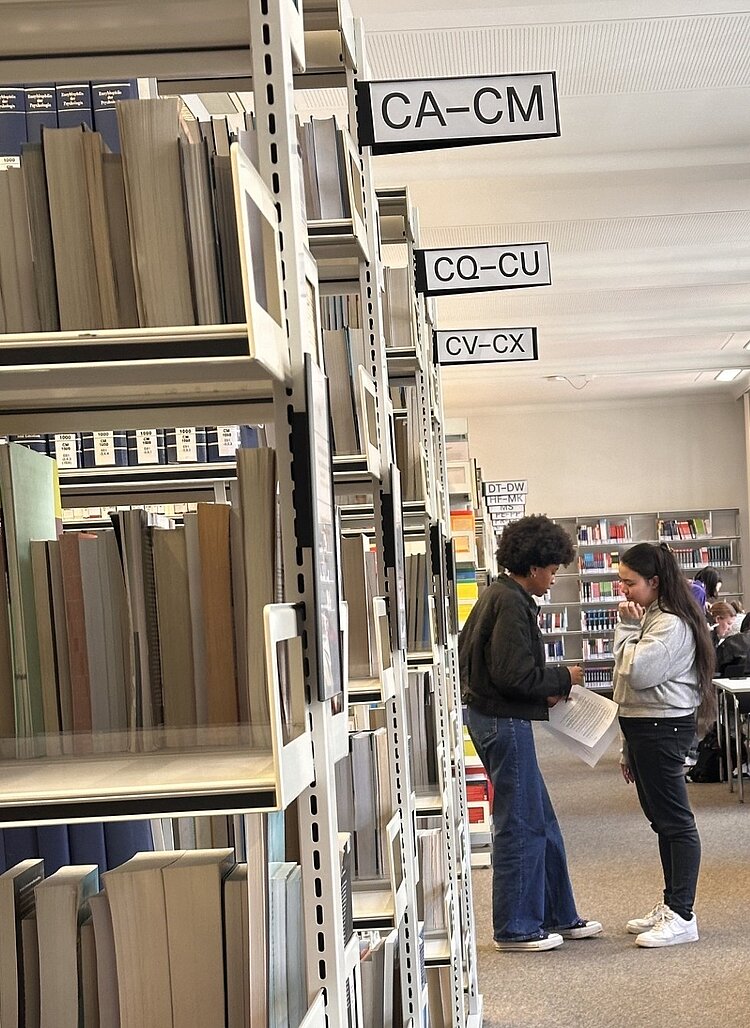 Zwei Studierende stehen zwischen Bücherregalen in einer Bibliothek und besprechen Unterlagen; im Hintergrund lernen weitere Personen an Tischen.