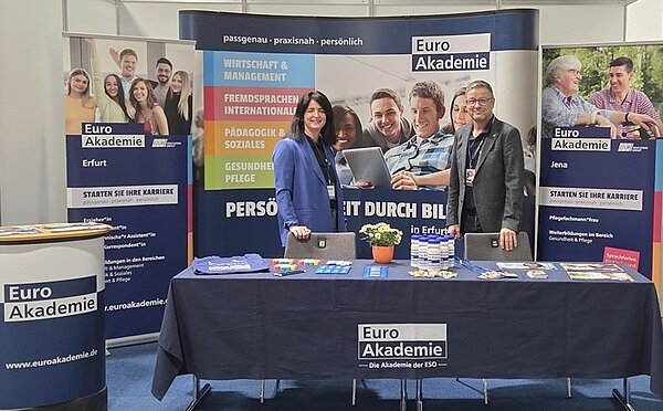 Euro Akademie Erfurt auf der JOBfinder Erfurt Schulleiter Uwe Schöneck von der Euro Akademie Erfurt steht mit einer Mitarbeiterin, beide in einem schicken Anzug, am Messestand der Euro Akademie, im Hintergrund große Roll-ups, ein Beratungs-Stehtisch links, ein langer Tisch, auf dem eine marineblaue Euro Akademie-Tischdecke liegt, auf der wiederum reichlich Informationsbroschüren und Flyer für die Messebesucherinnen und -Besucher ausliegen.