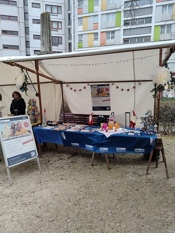 Überdachter Infostand der Euro Akademie auf einem Platz zwischen Hochhäusern; auf dem blau gedeckten Tisch liegen Flyer und kleine Bastelarbeiten, im Hintergrund hängen Lichterketten und ein großes Plakat „Genau meine Ausbildung“.