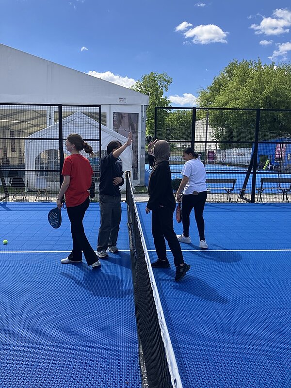 Vier Auszubildende der Euro Akademie stehen auf einem blauen Padelplatz, getrennt durch ein Netz. Zwei Azubis auf der linken Seite tragen Sportkleidung und halten Schläger in der Hand. Eine Person hebt den Arm, als würde sie abklatschen. Auf der rechten Seite stehen zwei weitere Personen, ebenfalls mit Schlägern. Im Hintergrund sind ein weißes Zelt, Bänke und grüne Bäume zu sehen. Der Himmel ist blau mit einigen weißen Wolken.