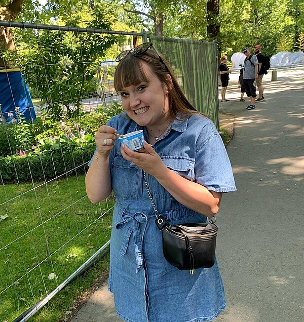 Eine junge Frau mit blauem Oberteilt, die ein Eis aus einem Becher isst und in die Kamera lächelt