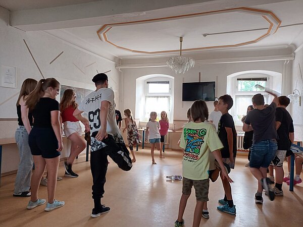 Sprachprojekte mit internationalem Flair Jungen und Mädchen stehen in einem Kreis in einer kleinen Turnhalle und machen auf einem Parkettboden Dehnungsübungen mit ihren Beinen