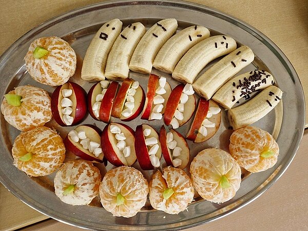 Gesund und gruselig: Kitaessen zu Halloween Auf einem silbernen Servierteller liegen gepellte Mandarinen die wir Kürbisse aussehen, Apfelspalten mit kleinen Marshmellows als Zähne und gepellte, halbierte Bananen, die mit Schokostreuseln zu Geistern verziert sind.
