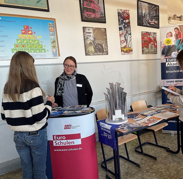 Eine Informationsveranstaltung in einem Raum mit mehreren Postern an der Wand. Im Vordergrund steht eine Frau an einem runden Infostand mit der Aufschrift 'Euro Schulen' und der Website www.euro-schulen.de. Sie trägt eine Brille, ein graues Tuch und ein Namensschild und gibt einer jungen Frau mit langem, blondem Haar und einem schwarz-weißen Pullover Informationen. Auf dem Tisch liegen Broschüren und Informationsmaterialien. Im Hintergrund sind weitere Poster und ein Stand mit der Aufschrift 'Euro Akademie' zu sehen.