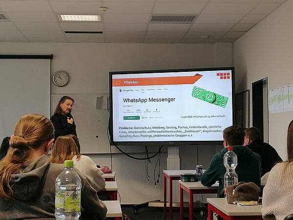 MeKoSax-Projekt „Trickfilmproduktion“ Eine Frau präsentiert verschiedene Plakate auf einem Bildschirm und davor sitzen mehrere Schüler in Sitzreihen.