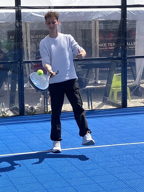 Ein junger Auszubildender mit hellem, langärmligem Oberteil und dunkler Hose steht auf einem blauen Tennisplatz. Er hält einen Tennisschläger in der rechten Hand und schlägt einen gelben Tennisball. Im Hintergrund sind Glaswände und unscharfe Schriftzüge auf Werbebannern sowie einige gelbe Stühle zu sehen. Die Szene ist sonnig, der Schatten des Spielers ist auf dem Boden erkennbar.