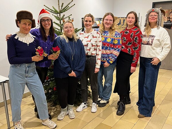 Sieben Schülerinnen posieren für ein Foto vor dem Weihnachtsbaum und halten zwei Weihnachtssterne in die Kamera. Einige tragen eine Weihnachtspullover, ein Mädchen hat eine Nikolausmütze auf dem Kopf.