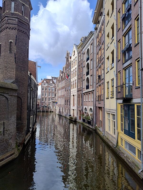 Ein Blick auf einen Kanal, der zwischen hohen, historischen Gebäuden in Amsterdam fließt. Die Häuser sind typisch für die niederländische Architektur, die sich der Himmel spiegelt sich im Wasser spiegeln.
