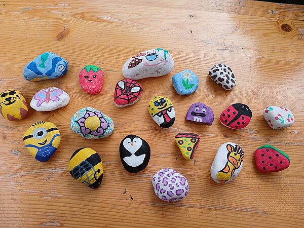 Eine Sammlung von bemalten Steinen mit verschiedenen Motiven wie Minions, Spiderman, Marienkäfer, Giraffe, Pinguin, Pizza und Erdbeeren, die auf einem Holztisch liegen.