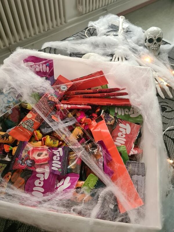 In einer großen Kiste sind viele Süßigkeiten für Halloween. Die Kiste und der Tisch sind mit Deko-Spinnweben, Deko-Totenköpfen und Deko-Knochen dekoriert.