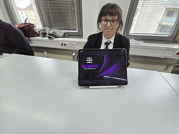 Eine Auszubildende im Anzug sitzt an einem Tisch in einem hellen Raum mit großen Fenstern. Vor ihr steht ein Tablet, dessen Bildschirm eine Präsentationsfolie mit dem Titel 'BALANCE' und einer stilisierten Waage zeigt.