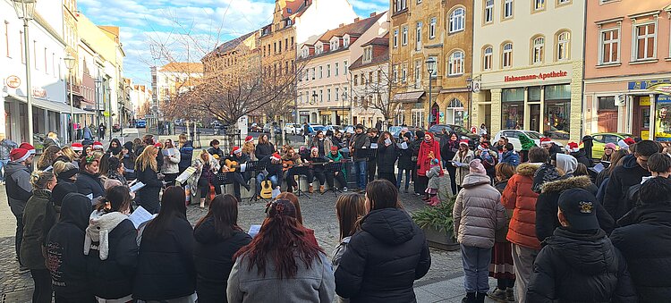 Zahlreiche Schüler*innen und Lehrkräfte stehen mit Winterjacken und Nikolausmützen in einer Fußgängerzone im Kreis, in der Mitte sitzen Musiker*innen mit Gitarren – gemeinsames Weihnachtssingen in der Innenstadt.