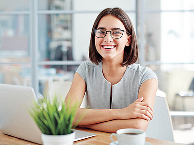 Auf dem Bild sitzt eine Frau an einem Tisch mit einem Laptop, einer Tasse Kaffee und einer Pflanze.