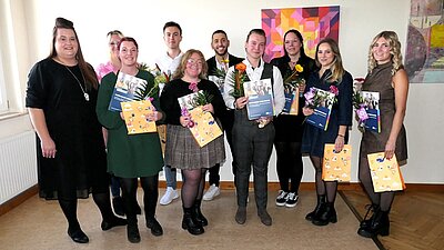 Die zehn Absolventinnen und Absolventen der Klasse EZ 21b posieren, mit Abschlusszeugnis und roten und gelben Gerbererblumen in der Hand, für ein gemeinsames Erinnerungsfoto.