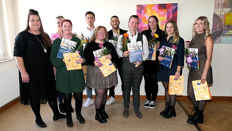 Die zehn Absolventinnen und Absolventen der Klasse EZ 21b posieren, mit Abschlusszeugnis und roten und gelben Gerbererblumen in der Hand, für ein gemeinsames Erinnerungsfoto.