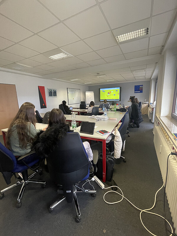 Seminarraum mit mehreren Teilnehmenden an Tischen; Laptops und Getränke stehen bereit, vorne ist ein großer Bildschirm mit einer Präsentation zu sehen.