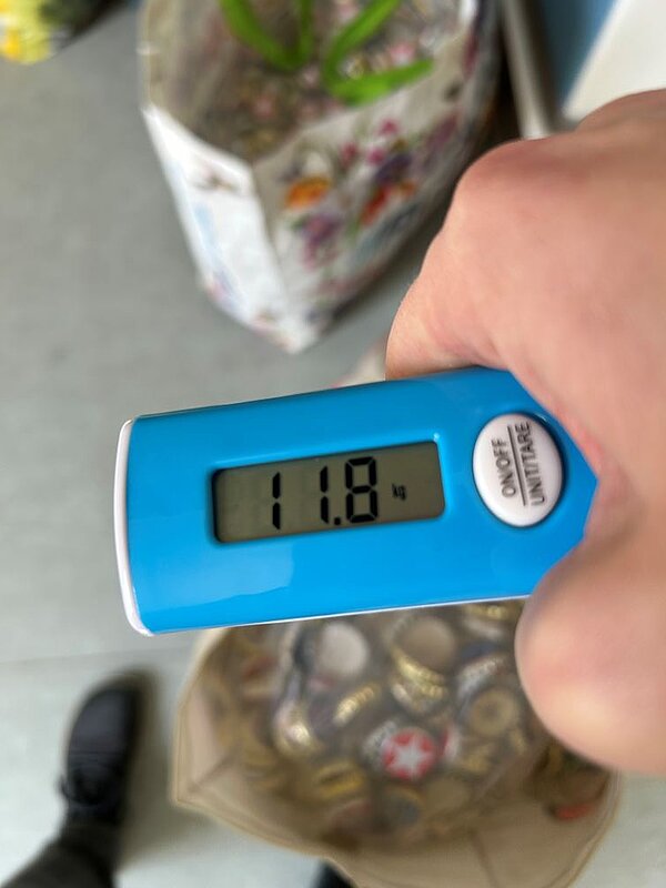 Zweite Wiegeaktion mit starkem Ergebnis Eine Hand hält eine digitale Hängewaage, die ein Gewicht von 11,8 kg anzeigt. Darunter ist eine durchsichtige Tasche mit gesammelten Kronkorken zu sehen. Im Hintergrund steht eine weitere, bunte Tasche mit weiteren gesammelten Korken.
