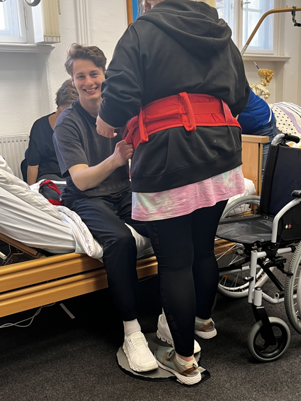 Eine Person sitzt auf einem Pflegebett und trägt dunkle Kleidung. Eine weitere Person steht davor, trägt eine schwarze Kapuzenjacke, eine rosafarbene Tunika, schwarze Leggings und weiße Turnschuhe. Sie legt der sitzenden Person einen roten Gurt um. Rechts steht ein Rollstuhl neben dem Bett. Im Hintergrund sind weitere Personen und ein Fenster mit weißen Vorhängen zu sehen.