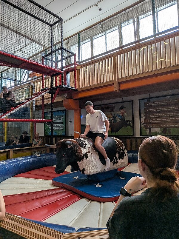 Ein junger Mann sitzt auf einem mechanischen Rodeo-Bullen mit schwarz-weißem Fell. Das Luftkissen darunter hat rot-weiße Streifen und blaue Flächen mit weißen Sternen. Im Vordergrund ist eine Zuschauerin zu sehen. Im Hintergrund sind Klettergerüste und Netze zu sehen, in denen weitere junge Menschen sitzen.