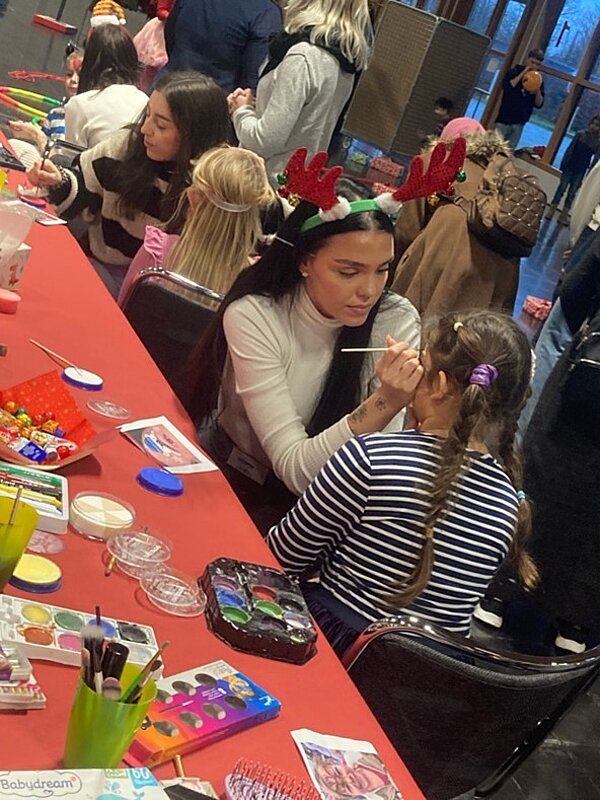 Eine Frau malt einem kleinen Mädchen das Gesicht an. Sie sitzen an einem Tisch mit verschiedenen Schminkutensilien und Bastelmaterialien. Andere Kinder sind im Hintergrund zu sehen, und die Szene scheint Teil einer Veranstaltung oder eines Festes zu sein.