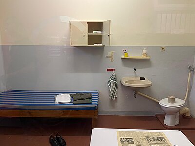 Nachgestellte Gefängniszelle im Kaßberg-Gefängnis mit schmalem Bett, kleinem Tisch, Waschbecken, Toilette, Regal mit Hygieneartikeln und gefalteter Kleidung.