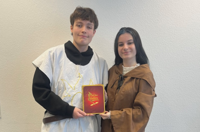 Die beiden Auszubildenden zu Kinderpfleger*innen stehen nebeneinander vor einer hellen Wand. Beide tragen mittelalterlich inspirierte Gewänder, eine Person ein weißes Oberteil mit gelbem Symbol, die andere einen braunen Umhang mit Kapuze. Sie halten gemeinsam ein rotes Buch mit goldener Schrift und Illustration, auf dem deutlich 'Bilder der Helden' zu lesen ist. Das Buch steht im Mittelpunkt der Szene und wird von beiden präsentiert.