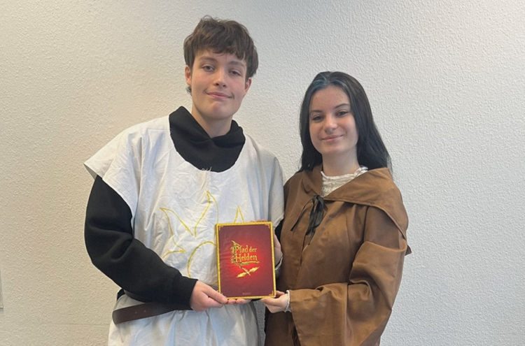 Die beiden Auszubildenden zu Kinderpfleger*innen stehen nebeneinander vor einer hellen Wand. Beide tragen mittelalterlich inspirierte Gewänder, eine Person ein weißes Oberteil mit gelbem Symbol, die andere einen braunen Umhang mit Kapuze. Sie halten gemeinsam ein rotes Buch mit goldener Schrift und Illustration, auf dem deutlich 'Bilder der Helden' zu lesen ist. Das Buch steht im Mittelpunkt der Szene und wird von beiden präsentiert.