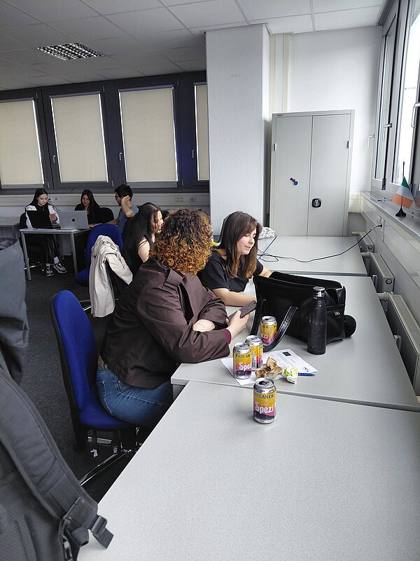 In einem Klassenzimmer sitzen mehrere Personen mit Laptops und Handys und besprechen sich.