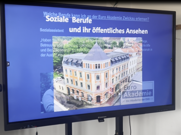 Ein großer Bildschirm zeigt eine Präsentationsfolie mit dem Titel 'Welche Berufe kann ich an der Euro Akademie Zwickau erlernen? Soziale Berufe und ihr öffentliches Ansehen'. Darunter ist der Begriff 'Sozialassistent' zu lesen. Auf dem Bildschirm ist ein halbtransparentes Bild eines historischen, mehrstöckigen Gebäudes mit einem spitzen Turm und gelb-beiger Fassade eingeblendet. Das Gebäude steht an einer Straßenecke und ist von Bäumen umgeben. Unten rechts auf dem Bildschirm ist das Logo der Euro Akademie mit dem Schriftzug 'Die Akademie der ESO' zu sehen. Die Präsentation findet im Rahmen des Tages der offenen Tür an der Euro Akademie Zwickau statt.