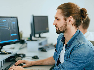Eine Person mit einem Dutt und Bart sitzt an einem Schreibtisch und arbeitet an einem Computer. Sie trägt ein blaues Hemd und konzentriert sich auf den Bildschirm, während sie auf der Tastatur tippt. Im Hintergrund sind weitere Monitore und Bürogegenstände zu sehen.