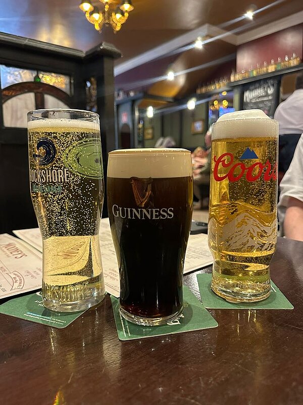 Drei gefüllte Biergläser auf einem Holztisch in einem traditionellen Pub. Von links nach rechts: ein helles Bier (Rockshore), ein dunkles Guinness und ein helles Bier (Coors). Im Hintergrund sind weitere Gäste und die gemütliche Atmosphäre des Pubs zu erkennen.