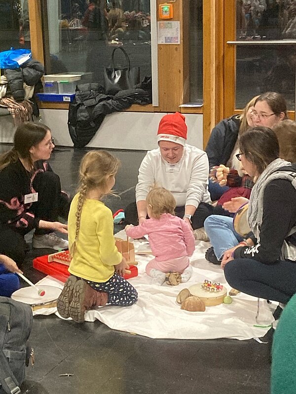 Eine Gruppe von Erwachsenen und Kindern sitzt auf dem Boden und nimmt an einer gemeinsamen Aktivität teil. Einige der Erwachsenen tragen Namensschilder und eine Person eine Nikolausmütze, und es sind verschiedene Musikinstrumente auf einer Decke verteilt. Die Szene spielt sich in einem Innenraum mit großen Fenstern im Hintergrund ab.